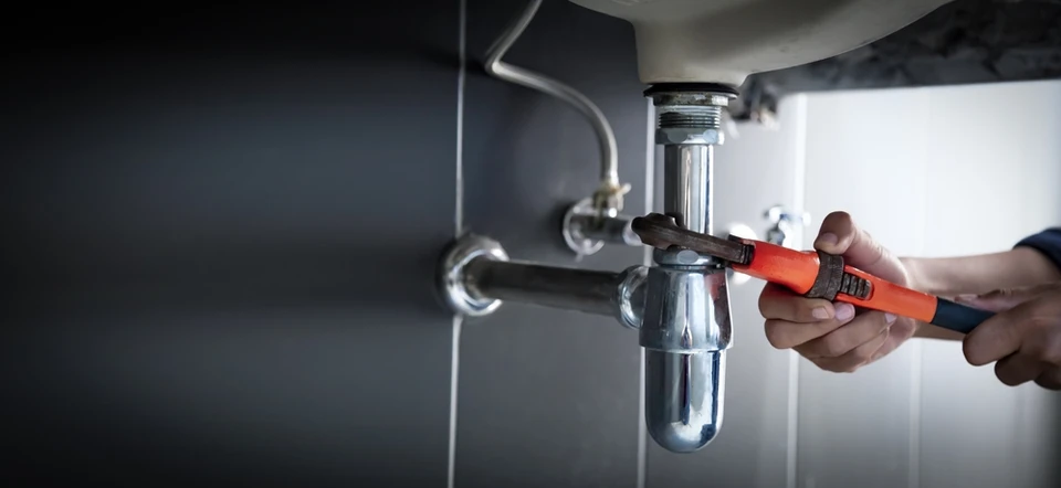 Plombier travaillant sous un évier de cuisine, utilisant une clé à molette orange pour serrer un siphon chromé