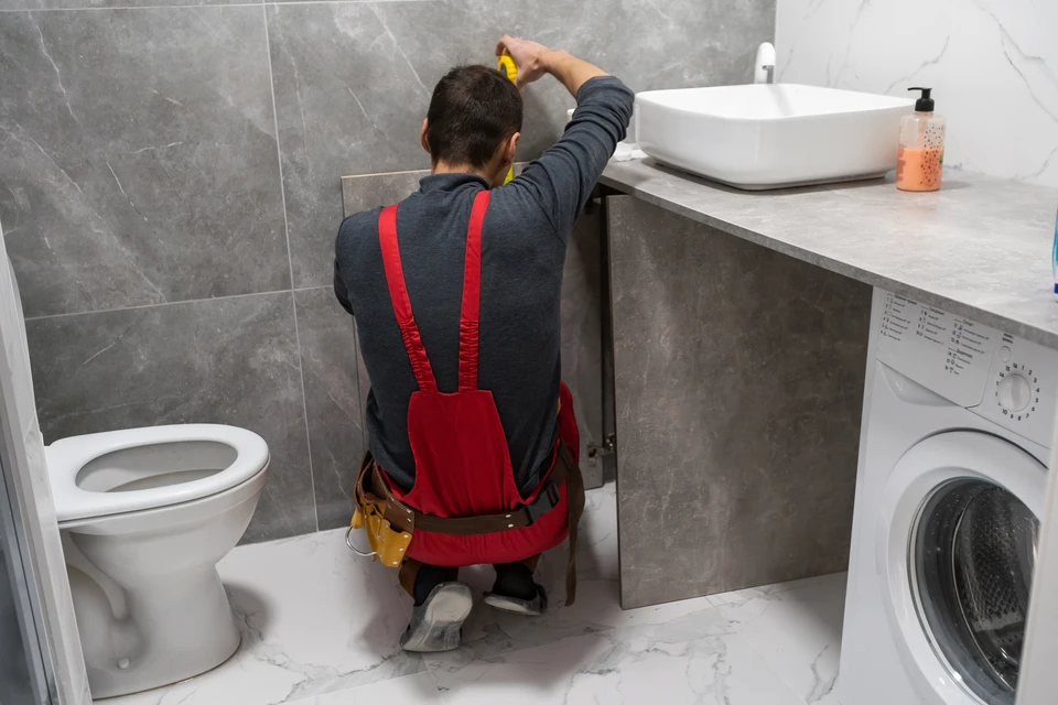 Plombier en salopette rouge travaillant dans une salle de bain moderne, agenouillé près d'un meuble vasque gris