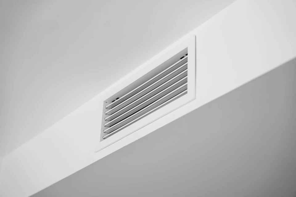 Homme barbu en t-shirt noir installant ou réparant un système de ventilation intérieur, travaillant sur un conduit d'aération circulaire fixé au plafond dans une pièce aux murs jaunes avec moulures décoratives