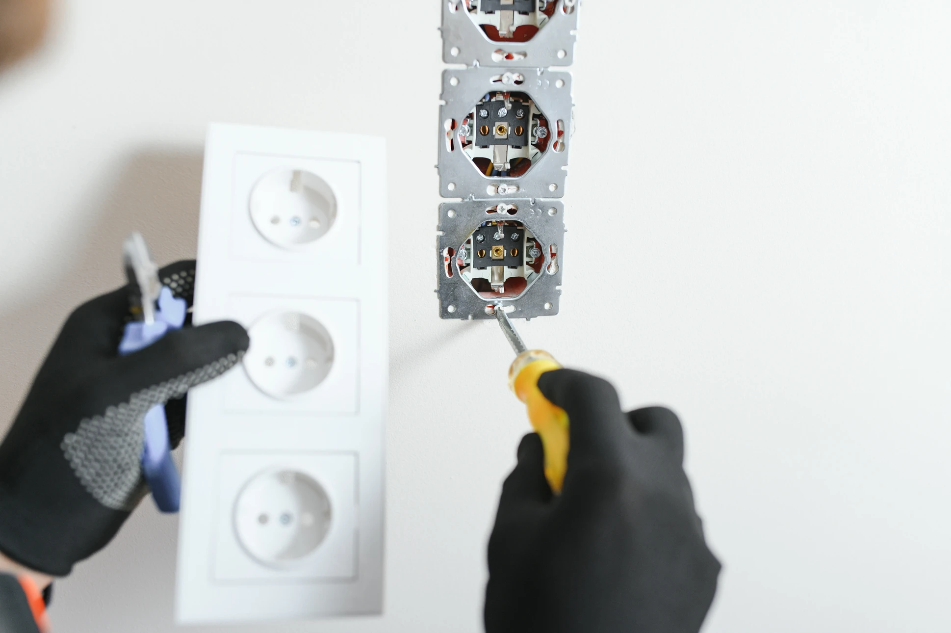 Électricien professionnel en chemise à carreaux travaillant sur installation électrique résidentielle, manipulant fils et connexions dans boîtier mural blanc