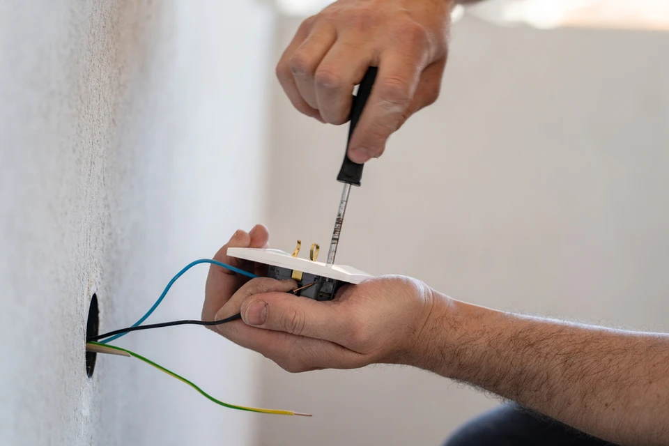 Photographie d'un électricien qualifié installant ou vérifiant un tableau électrique moderne avec disjoncteurs et protections différentielles dans un intérieur résidentiel. L'électricien, vu partiellement, effectue son travail sur le tableau ouvert, montrant des raccordements professionnels. L'arrière-plan montre un mur blanc ou neutre, évoquant un chantier de rénovation en cours. L'image doit transmettre la confiance, la professionnalité et la conformité des installations.