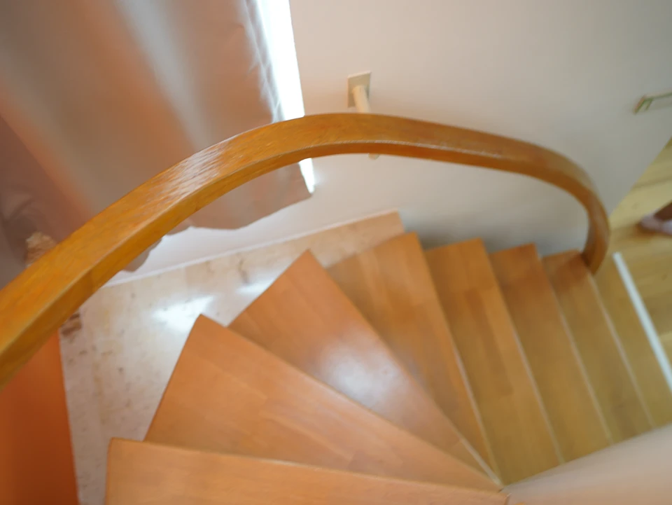 Photo d'un artisan en tenue de protection légère (gilet, gants, lunettes de sécurité) effectuant un aérogommage précis sur un escalier ou une poutre en bois clair en intérieur. La scène montre l'outil de projection fine en action, avec un focus sur la délicatesse de la technique et la clarté du matériau préservé après intervention. L'atmosphère doit être celle d'un chantier de rénovation haut de gamme, propre et organisé, reflétant le professionnalisme et le soin de CNR.
