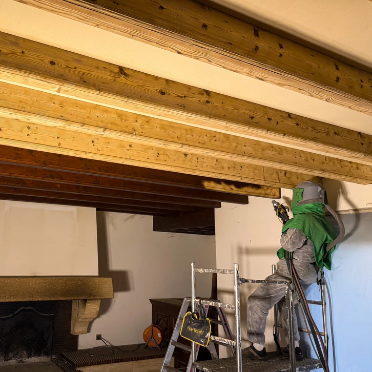 Photographie d'un artisan professionnel dans une maison en rénovation, discutant avec un couple de clients dans une pièce partiellement rénovée. L'artisan est en tenue de travail professionnelle, tenant un carnet de notes ou un tablet, souriant et établissant un contact visuel confiant. Le contexte montre clairement un chantier de rénovation intérieure (outils, matériaux, pièce transformée en arrière-plan), illustrant le rôle d'interlocuteur unique coordonnant la relation client du début à la fin.